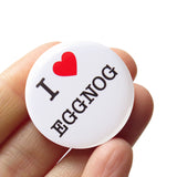 Round, white pinback button that says I LOVE EGGNOG in classic black text with "love" being represented by a red heart. Button is held in a hand.