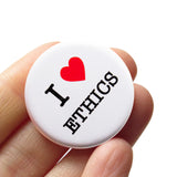 Round white 1.25" pinback button that reads I Love Ethics, love being a red heart. Badge held in a hand