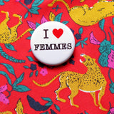 A round white pinback button that reads I LOVE FEMMES in a black serif font, the love being indicated by a red heart. Button is on red silk shirt patterned with leopards and florals.