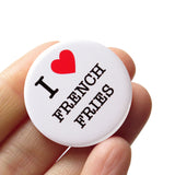 A round white button that reads I LOVE FRENCH FRIES. Love being represented by a red heart. Button is held in a hand.