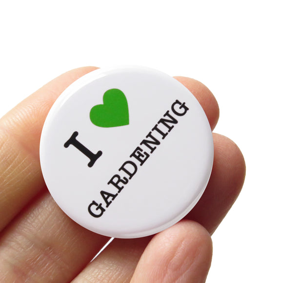 A white pinback button that reads I LOVE GARDENING, with love being represented by a green heart. Badge is held in a hand.