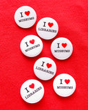 Seven round white 1.25" pinback button that read I LOVE LIBRARIES & I LOVE MUSEUMS, love being represented by a red heart. Buttons are pinned to a red sweatshirt.