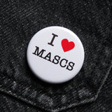 A round white pinback button that reads I LOVE MASCS in a black serif font, the love being indicated by a red heart. Button is pinned to a black denim pocket