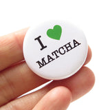 A round white pinback button that reads I LOVE MATCHA, with a green heart representing love, and the rest of the phrase is in a classic serif font. Button held in a hand.
