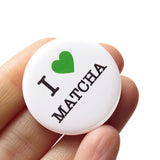 A round white pinback button that reads I LOVE MATCHA, with a green heart representing love, and the rest of the phrase is in a classic serif font. Button held in a hand.