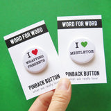 Two round white pinback buttons that read I LOVE WRAPPING CHRISTMAS PRESENTS with love being a red heart & the other button reading I LOVE MISTLETOE, "love being represented by a green heart.  Buttons are held in a hand, on WORD FOR WORD branded backing cards for What We Really Love