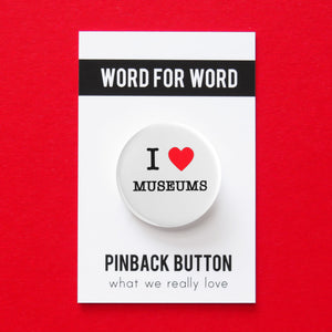 A round white pinback button that reads I LOVE MUSEUMS, love being represented by a red heart. Button is held in a hand.