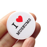 A round white pinback button that reads I LOVE MUSEUMS, love being represented by a red heart. Button is held in a hand.