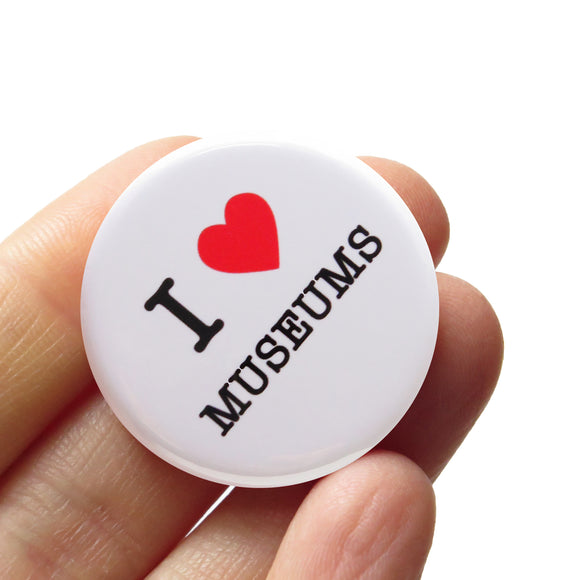 A round white pinback button that reads I LOVE MUSEUMS, love being represented by a red heart. Button is held in a hand.