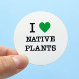 A round white sticker held in the corner with a thumb & forefinger against a light blue background. Sticker reads: I LOVE NATIVE PLANTS with love being represented by a green heart.