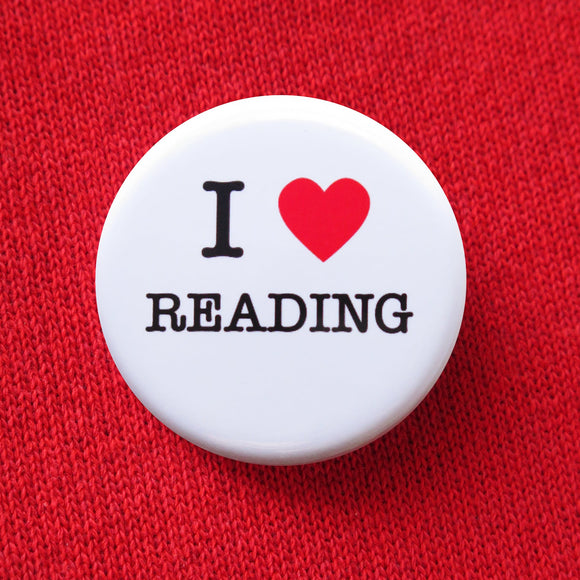 A round white pinback button that reads I LOVE READING, love being represented by a red heart. Button is held in a hand.