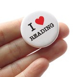 A round white pinback button that reads I LOVE READING, love being represented by a red heart. Button is held in a hand.