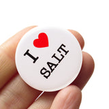 A round white button that reads I LOVE SALT. Love being represented by a red heart. Button is held in a hand.