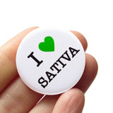 Round pinback button on a white background that says I LOVE SATIVA. Love is depicted by a green heart.  The other text is black. Button is held in a hand.