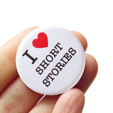 Round white pinback button that reads I heart short stories with the heart being a bright red heart. Button is held in a hand.