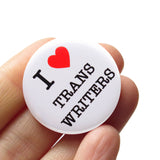 Round white 1.25" pinback button with a bright read heart that reads I HEART TRANS WRITERS in classic black type. Button is held in a hand