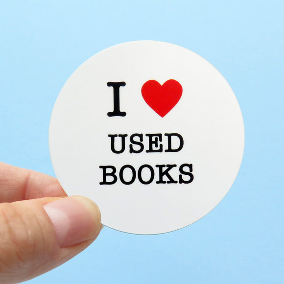 A round white sticker held in the corner with a thumb & forefinger against a light blue background. Sticker reads: I LOVE USED BOOKS with love being represented by a red heart.