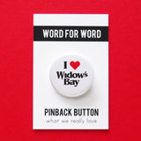 A round white pinback button that reads I LOVE WIDOWs BAY, love being represented by a red heart. Button is pinned to a Word For Word, What We Really Love branded backing card.Setting on a red table.
