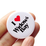 A round white pinback button that reads I LOVE WIDOWs BAY, love being represented by a red heart. Button is held in a hand.