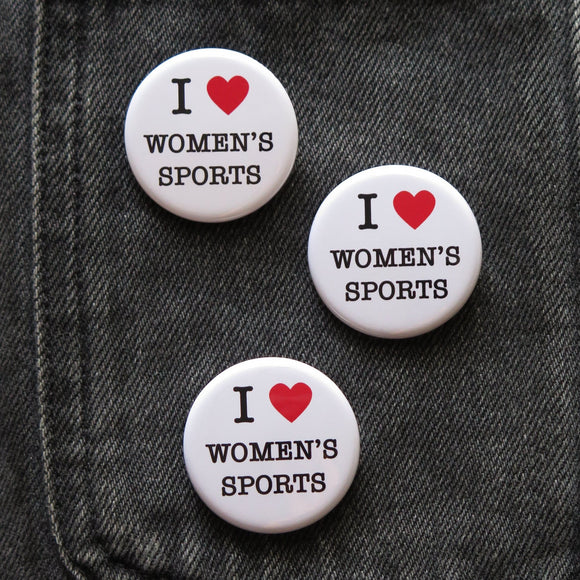 Three round white pinback buttons that reads I LOVE WOMENS SPORTS with love been represented by a red heart. Buttons are pinned to a black denim pocket.