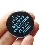 A round black pinback button that reads I STILL CHRISTINE BLASEY FORD in light blue text. Button is held in a hand.