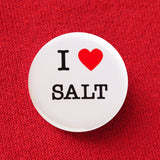 A round white button that reads I LOVE SALT. Love being represented by a red heart. Button is pinned to a red sweatshirt