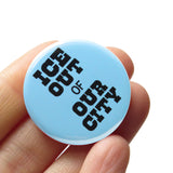 Round light blue pinback button that reads: ICE OUT of OUR CITY in chunky black text. Badge is held in a hand.