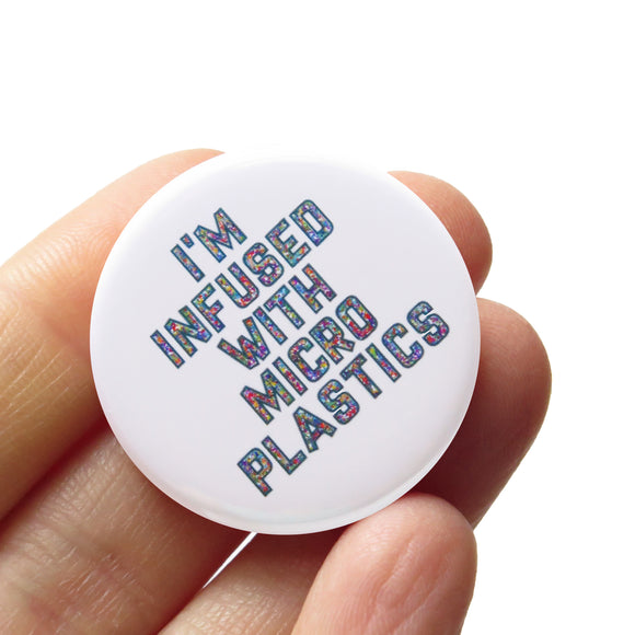 Round white pinback button that reads I'M INFUSED WITH MICROPLASTICS in a hollow font filled with tiny colored dots. Button is held in a hand.