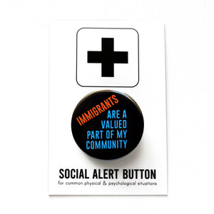 Round black pinback button that reads: IMMIGRANTS ARE A VALUED PART OF MY COMMUNITY in red-orange and blue text. Button is held in a hand.