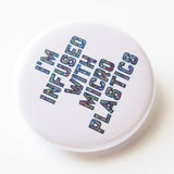 Close up photo of a round white pinback button that reads I'M INFUSED WITH MICROPLASTICS in a hollow font filled with tiny colored dots. 