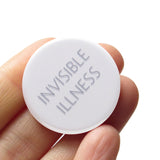 Round white 1.25" pinback button that reads INVISIBLE ILLNESS in thin gray-blue text. Button is held in a hand.