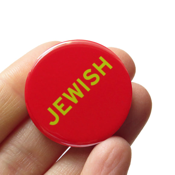 Round red pinback button reads JEWISh in light green text. Badge is held in a hand.