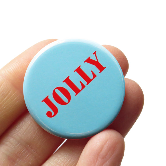 Round, pale blue-green pinback button that reads JOLLY in bright red text. Button is held in a hand