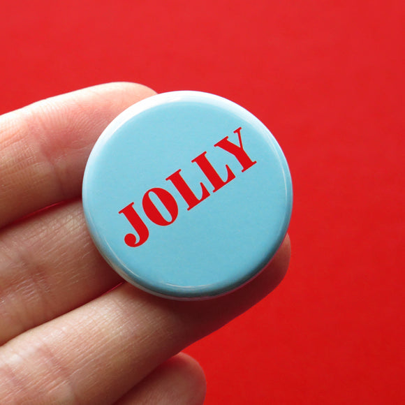 Round, pale blue-green pinback button that reads JOLLY in bright red text. Button is held in a hand in front of a background. 