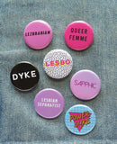 A multicolor group of gay and queer pinback buttons on faded blue denim. Button sayings include: Dyke, Lesbo, Queer Femme, Sapphic, Lezbrarian, Power Dyke & Lesbian Separatist.