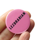 Round pink colored pinback button which reads LEZBRARIAN in a black italic sans serif font. The button is held in a hand.