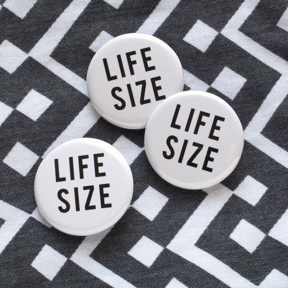 Three white round finback buttons that read LIFE SIZE in black text, on a patterned black and white denim background.