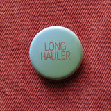A round pale blue green pinback button that reads LONG HAULER in dark orange thin text. Button is pinned to rust orange denim. For those suffering with long covid.