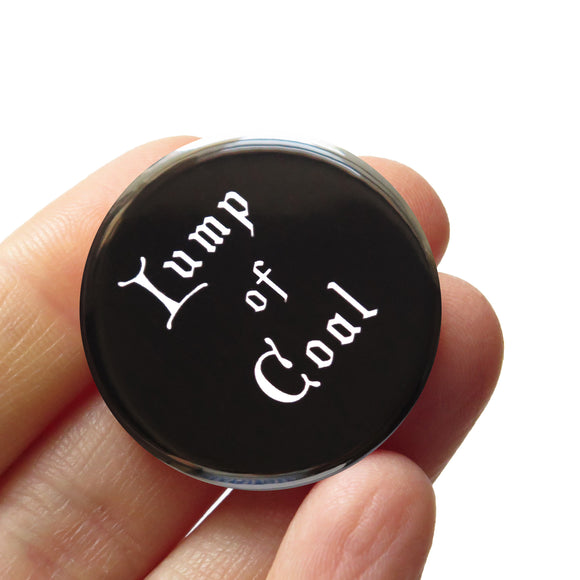 Black round pinback button that reads LUMP OF COAL in white old fashioned font. Badge held in a hand.