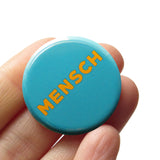 Round blue teal pinback button that reads MENSCH in yellow san serif font. Pinback button held in a hand.