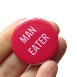 Round magenta pink pinback button that reads MAN EATER in a light pink san serif font. Pinback button is held in a hand.