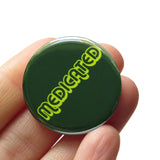 Round dark green button that reads MEDICATED in a neon green font. Badge is held in  a hand