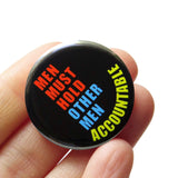 Round black pinback button that reads MEN MUST HOLD OTHER ACCOUNTABLE in neon yellow, orange and blue san serif text. The button is held in a hand.