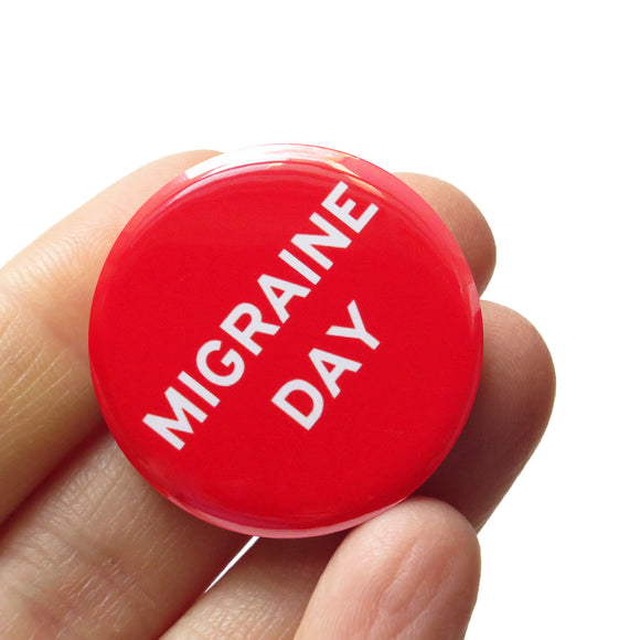 Shiny bright red pinback button with white san serif test reading, Migraine Day.  The button is held in a hand.