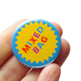 A round blue pinback button with a circular yellow starburst in the middle, and mixed color text that reads MIXED BAG. Badge is held in a hand.