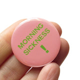 A round light pink pinback button that reads MORNIG SICKNESS ! in lime green text. Button is held in a hand.