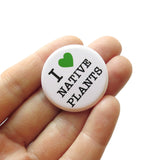 A white hand holds a  white pinback button that reads I LOVE NATIVE PLANTS, with love being represented by a green heart. 