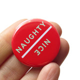 Round red 1.25" pinback button.  The button is holiday themed, with a red background and light mint green text that reads NICE in one direction and NAUGHTY flipped 180 degrees in the other direction, with a line diving the two words, and allowing for the button to be flipped for different readings.. Button is held in a hand.