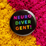 Round, black pinback button that reads NEURO-DIVER-GENT! In three lines with varied bright colors in an askew sans serif font. Button is pinned on a sweater