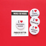 Four round white pinback I heart buttons that read:  I Love National Parks, I Love Vinyl Records, I LoveVintage, I Love French Fries. all buttons are on a red background, and one button is on a branded backing card.